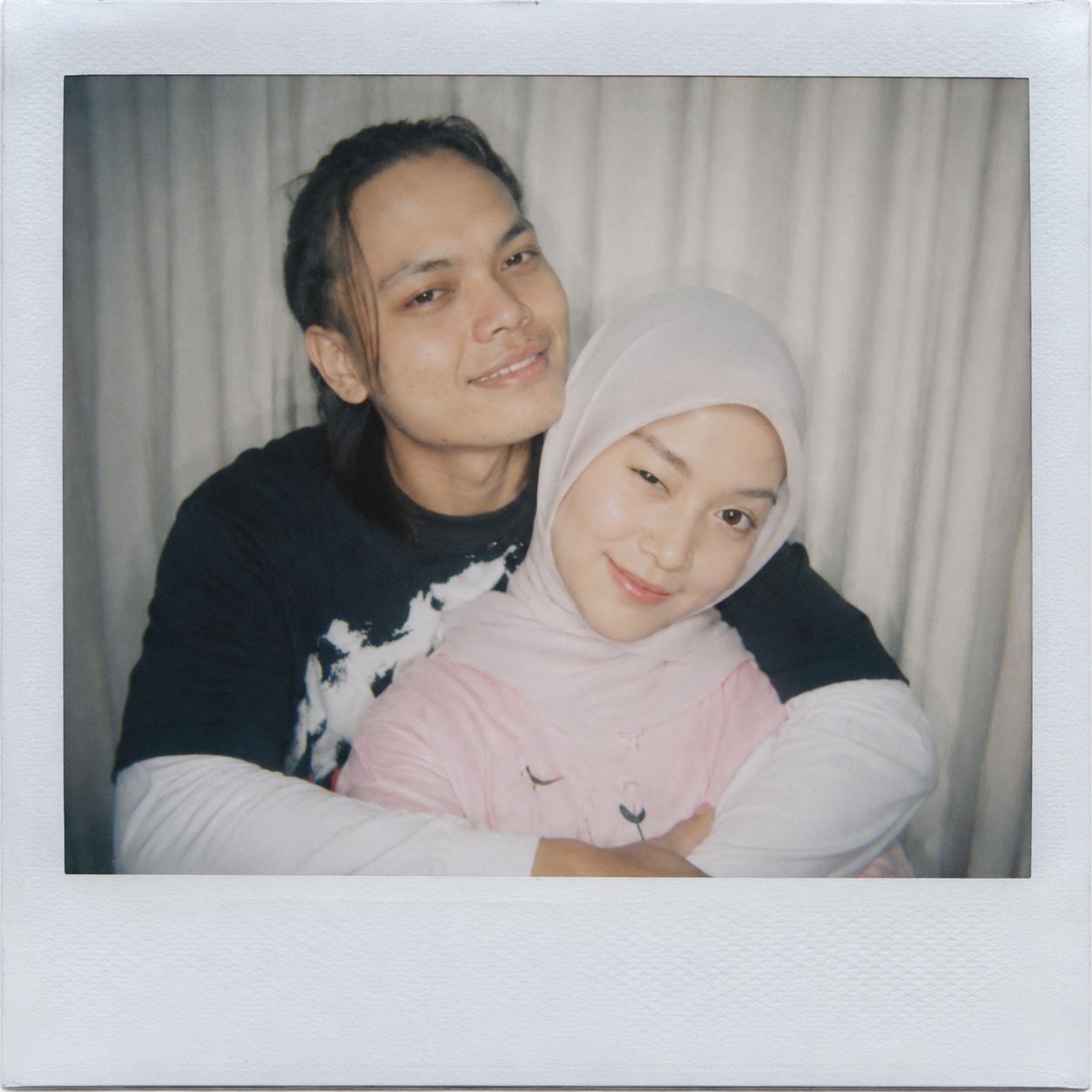 Create an image captured with a Polaroid camera. The photo should look like a casual snapshot, with no clear subject or staged props. Apply a slight blur effect and ensure consistent lighting, as if illuminated by a camera flash in a dark room, diffused across the entire image. Do not alter any faces. Replace the background behind the two people with white curtains. The man should be gently hugging the woman from behind, and both are standing close together smiling at the camera
