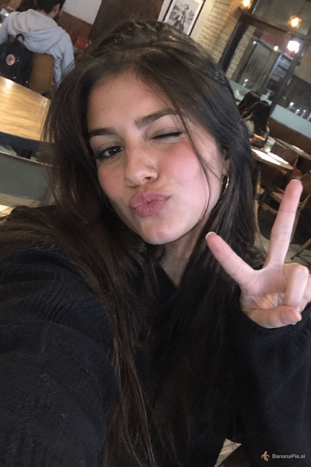 A young woman taking a selfie in a cafe. She's doing a cute expression with pursed lips, winking, one hand doing a piece sign. Irregular angle, amateur low quality photo
