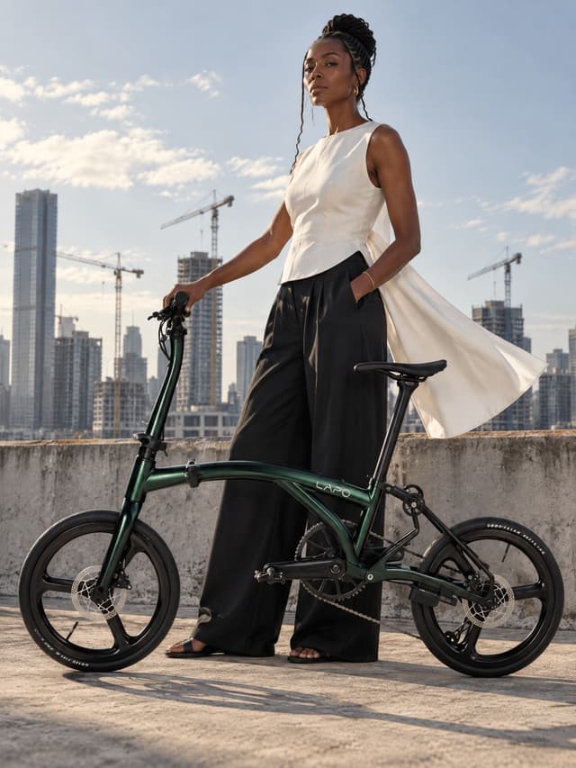 Create ONE vertical 3:4 premium fashion campaign photograph, not a collage, as a scene compositing transformation of the supplied bicycle product reference. Reference 1 defines the EXACT bicycle geometry and all component shapes; keep that geometry, stance and part layout unchanged. Reference 2 shows the same bicycle in the DEEP GREEN paint scheme and is used ONLY for that finish, its metallic depth and its headtube treatment; Reference 3 is the authoritative custom LAPO logo outline, only for small on-frame branding. One original fictional ADULT model, one unfolded bicycle. Preserve bicycle shape accurately: small equal wheels each THREE broad spokes, curved mainbeam, slender long seatpost and hollow saddle, tall curved stem, black fork, round rear folding joints, realistic connected drivetrain and chain. No generic diamond frame, extra tubes, wrong wheels, duplicated parts. Bike has both tires grounded and is stabilized at the explicitly described hand contact. No riding, pushing, lifting, floating, impossible balance. Black parts remain black; metallic short headtube only follows reference. Reproduce small custom LAPO outline on REAR half of mainbeam near seatpost, open stylized A and separated P stem, not a standard typed font. Full bike fits clearly and realistically in shot. Human face prominent and memorable, believable weight and fingers, couture editorial posture. Ground contact shadows physically agree with light. Person, fabric and bicycle remain separable, no garment inside wheels or chain. Visceral high-end fashion photograph, realistic pores, tactile materials, photographic lighting not plastic CGI, thoughtful installation-like visual event. No title, no large logo, no unrelated brand or watermark, no extra people. Paint scheme DEEP GREEN: deep forest-green metallic frame with a matching deep-green metallic short headtube shell, a dark chain and a small light wordmark on the mainbeam, metallic depth matched to Reference 1. Long stem, fork, seatpost, saddle, cranks and the three-broad-spoke wheels stay black. A fictional adult woman age 36, deep brown skin, braided hair gathered high, cool unimpressed gaze. Wearing a structured white sleeveless top with wide black trousers, flat sandals, a thin gold bangle. Pose: she stands very upright at the parapet with the bicycle held beside her, one hand on the grip, hem lifting in the wind. Set: the outer edge of a high roof with a construction-crane skyline compressed between her and the bicycle. Hard light from behind the camera compresses the crane skyline into the gap between the two silhouettes, and one corner of fabric swings toward the lens. Low camera slightly below the waist line, the grown model occupying about half of the frame with the complete unfolded bicycle fully visible across the lower half. The bicycle faces the LEFT side of the frame, seen from its DRIVE side, chainring, chain and rear derailleur toward camera. Any wordmark visible on the frame must read correctly left to right, never mirrored or reversed. Both tires ON the ground, the complete unfolded bicycle readable across the frame, no motion blur, one edited finished photograph.