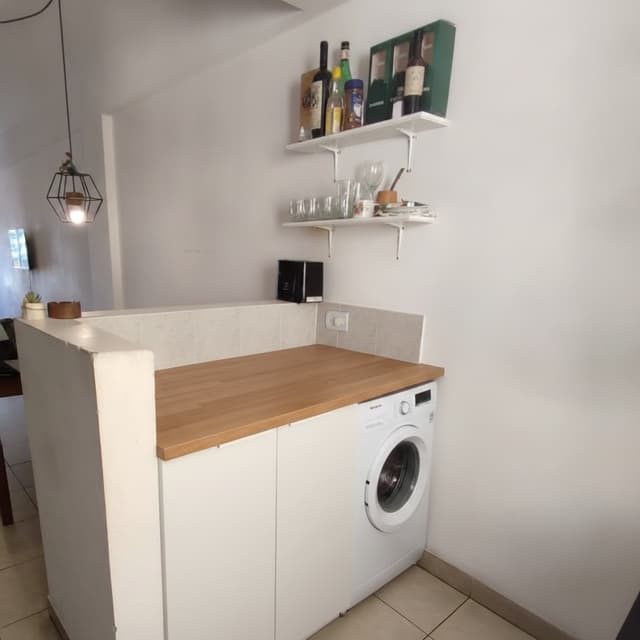 Lavadero de diseño tipo carpintería a medida, encimera de roble claro macizo de 4 cm de espesor, mueble blanco mate con sistema push-open, lavadora integrada bajo encimera a la derecha, almacenamiento oculto a la izquierda, estética minimalista escandinava, interiorismo moderno, fotografía hiperrealista, iluminación natural, no empty space, high-end custom cabinetry, luxury apartment laundry area.