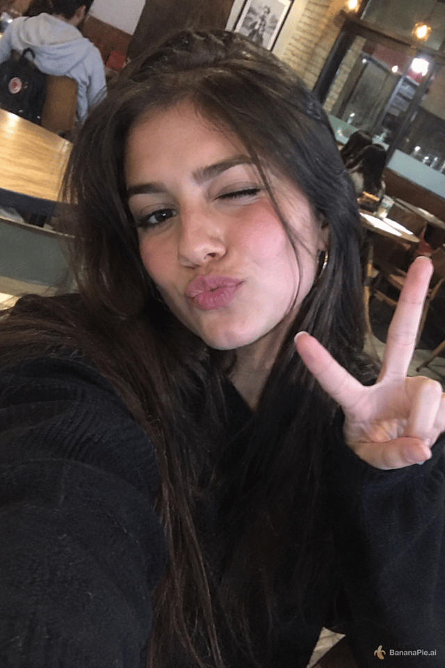 A young woman taking a selfie in a cafe. She's doing a cute expression with pursed lips, winking, one hand doing a piece sign. Irregular angle, amateur low quality photo