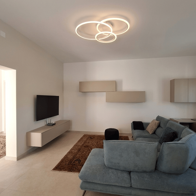 Crea un foto ritocco di questa foto del soggiorno, togliendo il mobiletto vecchio della tv, ed inserisci l'arredo come indicato nel disegno 2d dove sono indicati i tre prospetti della stanza con l'inserimento del mobilio. Nel lato tv viene posto un mobile sospeso, nella parete a fianco ci sono due pensili della stessa dimensione sfalsati tra di loro. Nella parete dietro al divano ci sono altri due pensili ed uno scrittoio, ma forse è fuori dal campo visivo. Pensili tinta beige. Lampadario come immagine allegata.