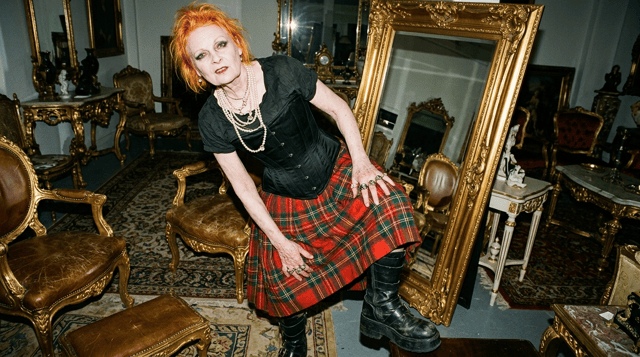 Subject: An elderly woman resembling Vivienne Westwood, with short electric orange hair, pale skin, and heavy dark eyeliner. She wears a thick tartan skirt, a tight black corset with visible boning, and multiple layers of pearl necklaces. She wears heavy black platform leather boots.
Environment: A luxurious rococo furniture showroom. A giant ornate gold-gilded standing mirror.scuffed leather, matte finish, heavy wear。accurate tartan weave, sharp pattern edges。weathered skin, visible veins, pale, realistic skin pores。
Camera & Lighting: Extreme low-angle shot, looking up at her. Harsh direct camera flash, 35mm documentary photography, high contrast, slight wide-angle distortion, raw film grain, unretouched skin texture, Juergen Teller style. --ar 16:9 --style raw --v 6.0