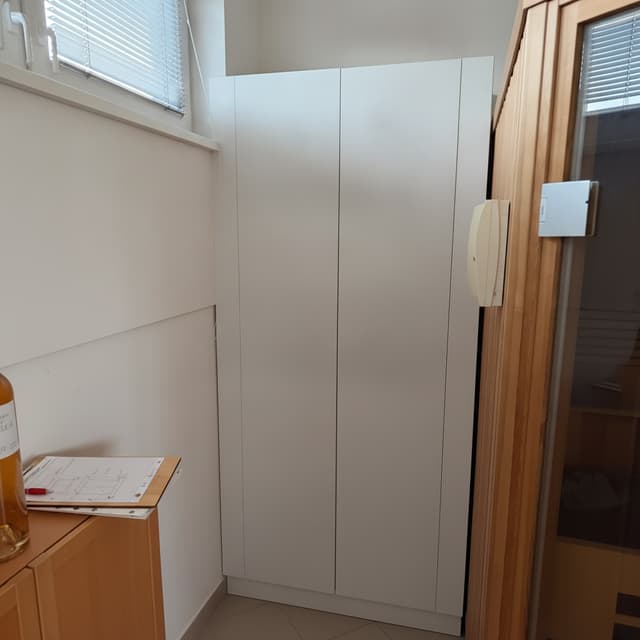 Image 1 is the base photo of a small storage niche. Image 2 is a hand-drawn front elevation sketch of the cabinet to install.

Edit image 1: remove all clutter from the niche (round table, chairs, bags, cooler box, boxes, mat, hammock chair). Keep everything else unchanged: white walls, the window with blinds, the sloped ceiling edge, the wooden sauna on the right, the wooden cabinet on the left foreground, tiled floor, lighting, perspective and camera angle.

Place a white cabinet in FRONT of the niche, not inside it. The cabinet stands at the front opening of the niche, between the wooden cabinet on the left and the sauna on the right. Its front closes off the niche, so the back wall of the niche is no longer visible. The cabinet is tall: its top edge reaches up to the bottom of the window (window sill height), so the window stays fully visible above it.

Follow the sketch:
- Total width 101 cm, total height 188 cm.
- Exactly two flat doors of equal width (about 43.5 cm each) with a thin vertical gap in the middle. No handles, no knobs, no visible hardware. Do not draw an X or diagonal lines.
- Narrow filler panel on the left (4 cm) and a wider one on the right (10 cm), both flush with the door fronts, no offset.
- A recessed plinth, 10 cm high, at the bottom across the full width.
- Matte white lacquer finish on all parts, even narrow joints, tight fit to the side walls and ceiling line.

Photorealistic, matching the existing daylight, soft shadows and color tone.